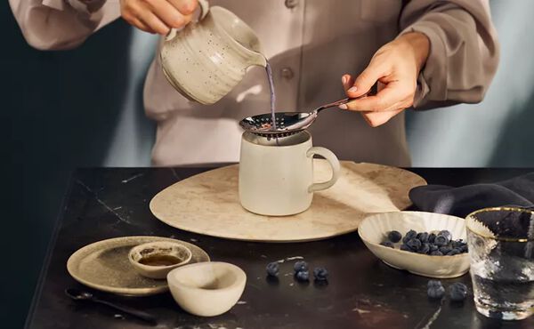 A person pours warm moon milk through a strainer into a mug, surrounded by blueberries and calming ingredients.