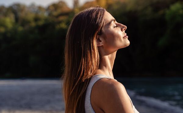 A woman with long brown hair stands outdoors with her eyes closed, facing the sunlight and appearing relaxed. She is wearing a sleeveless top and is surrounded by a natural setting with trees in the background and a body of water nearby.