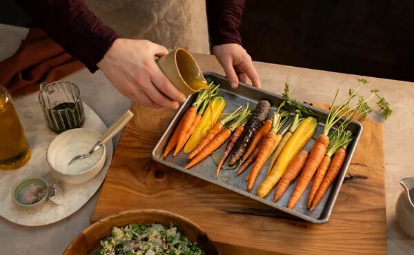 Person drizzling olive oil over a tray of colorful heirloom carrots on a wooden cutting board, surrounded by bowls of dressing, seasonings, and salad.