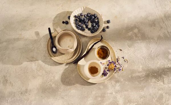 Ingredients for purple moon milk, including fresh blueberries, vanilla, cinnamon, and oat milk on a neutral-toned surface.