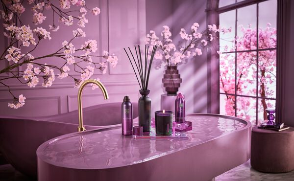 A purple bathroom with pink cherry blossom trees. There is a large purple sink with a gold tap. On the sink sits The Ritual of Yozakura products, the fragrance sticks, shower foam, candle and body lotion mousse.