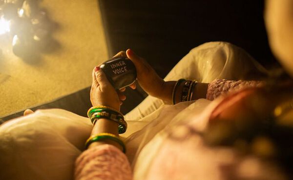 A close-up image from above of a woman sitting cross-legged wearing white trousers and a coral top with flower embroidery and green bangles. She is holding a smooth black stone in her two hands that says Inner Voice in white on it.
