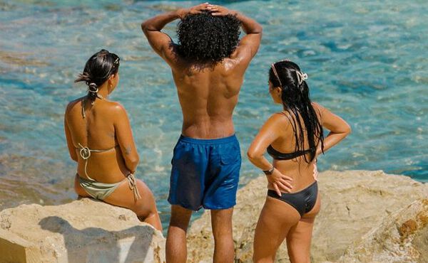 Three friends in swimwear relaxing by the sea, enjoying sunshine, connection and carefree summer moments together.