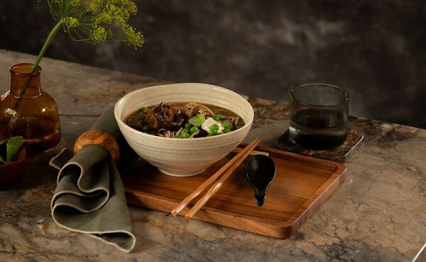 Mushroom miso broth: Bowl of mushroom miso soup with noodles, garnished with green peas and mushrooms, served on a wooden tray with chopsticks and a black spoon, next to a glass of water and a brown vase with green flowers on a marble countertop.
