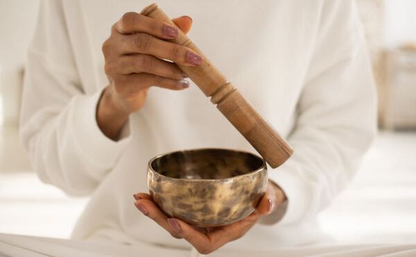 A woman dressed white – we don’t see her face but we can see she is sat cross-legged. She is holding a gold metal sound bowl and singing bowl stick that she is gliding on the edge of the bowl.