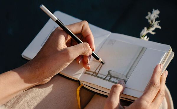 A woman who is sat down, she is wearing beige linen trousers. We don’t see her face, just her hands, she is drawing a chair and a bench with a pencil in a notebook.