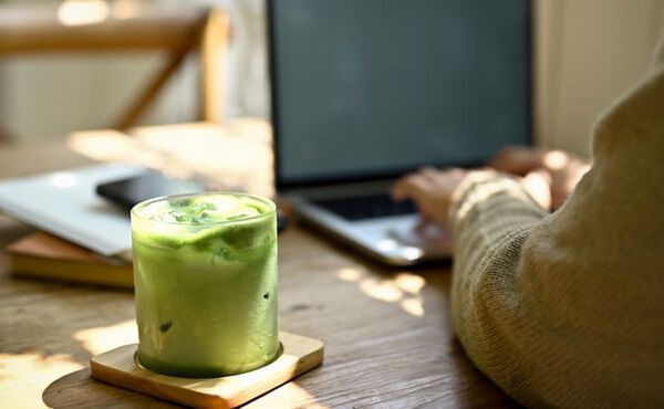 A person types on a laptop at a sunlit wooden table with a glass of iced matcha latte in the foreground, placed on a wooden coaster next to notebooks and a smartphone.